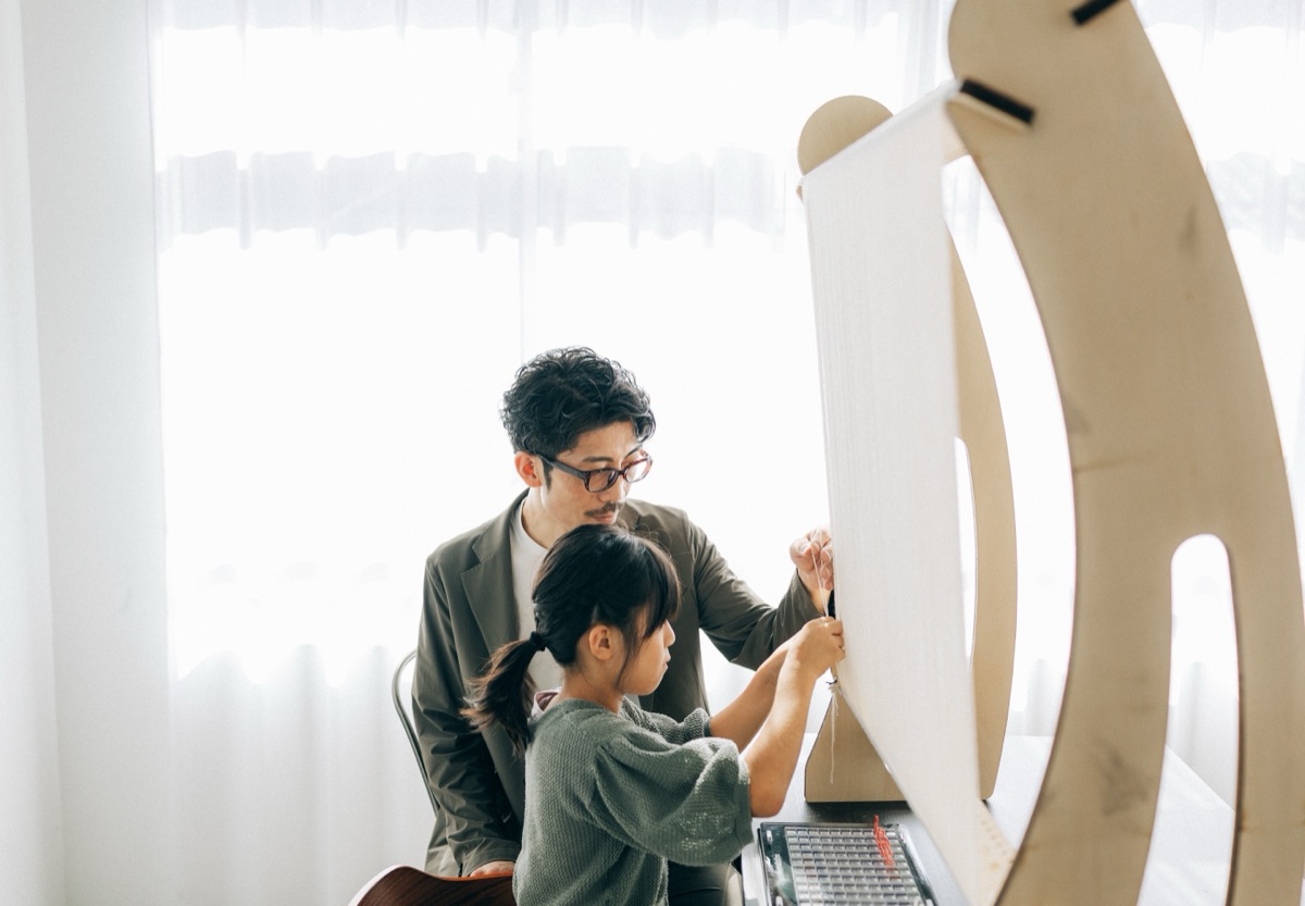 窓辺の織り機に向かう子どもと、そのうしろから手元を支える大人。