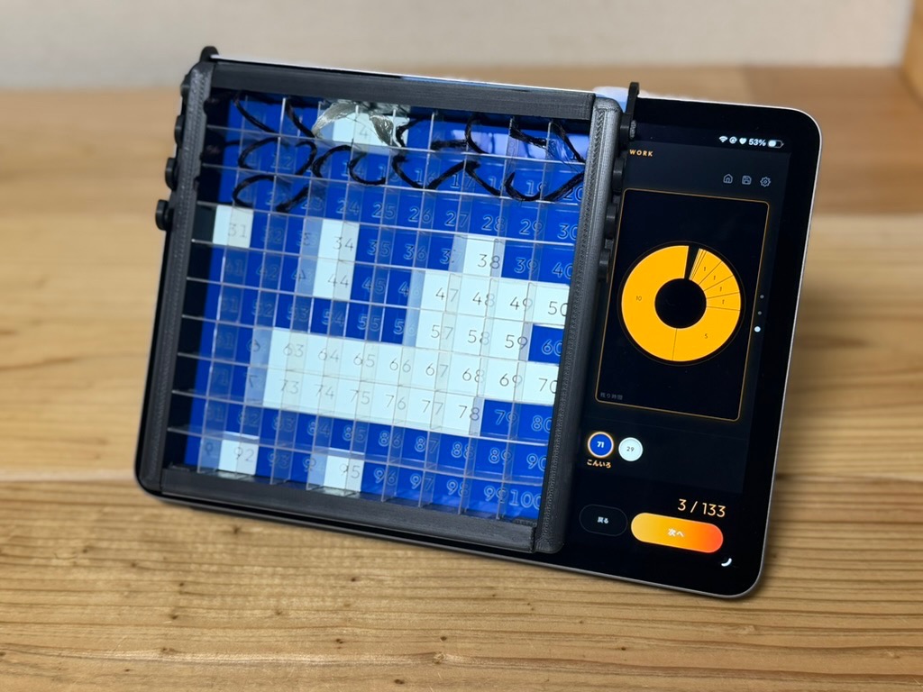 タブレットに 100 マスのパレットを重ねた制作モードの画面。右側に残り時間を示す輪と、次に使う糸の色が並ぶ。