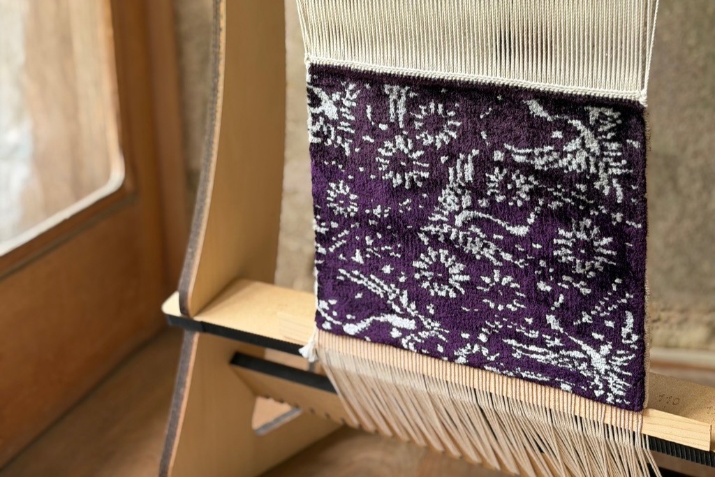 窓辺の織り機に張られた、織り上がった緞通。紫地に白い花の文様。